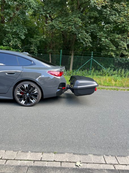 Grauer Sportwagen mit einem Anhänger am Heck, auf einer Straße neben Bäumen.