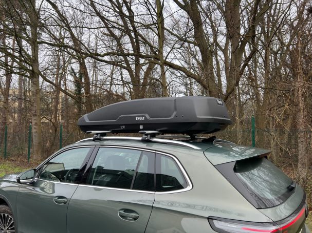 Dachbox auf einem grauen Auto, geparkt unter Bäumen.
