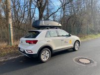 Ein weißer SUV mit Dachbox steht an einer Straße, umgeben von Bäumen.