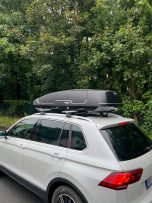 Ein weißer SUV mit einem schwarzen Dachgepäckträger auf einem Waldweg.