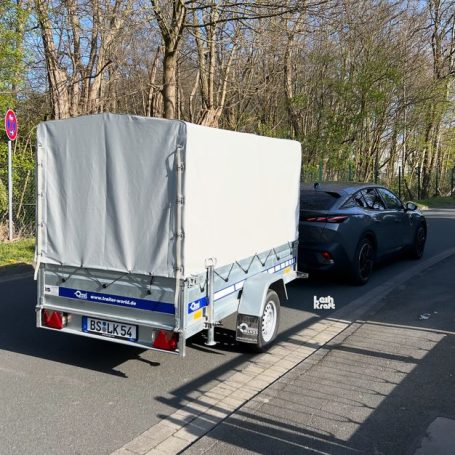 Ein PKW mit Anhänger auf einer Straße, umgeben von Bäumen und Verkehrszeichen.
