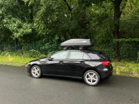 Schwarzes Auto mit Dachbox, parkend an einer Waldstraße.