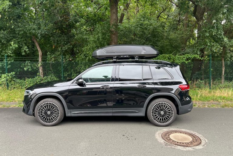 Schwarzes Auto mit Dachbox parkt unter Bäumen auf einer strahlenden Sonnenstraße.