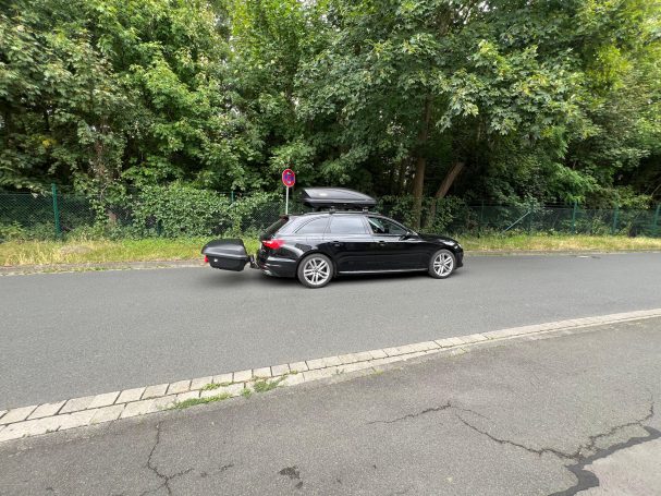 Schwarzes Auto mit Dachbox in einer ruhigen Straße, umgeben von Bäumen.