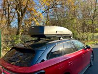 Roter Kombiwagen mit Dachbox, umgeben von Bäumen im herbstlichen Umfeld.