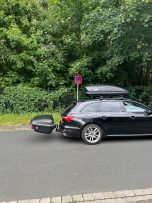 Schwarzes Auto mit Dachbox hält an einer Straße neben einem Parkverbotsschild.