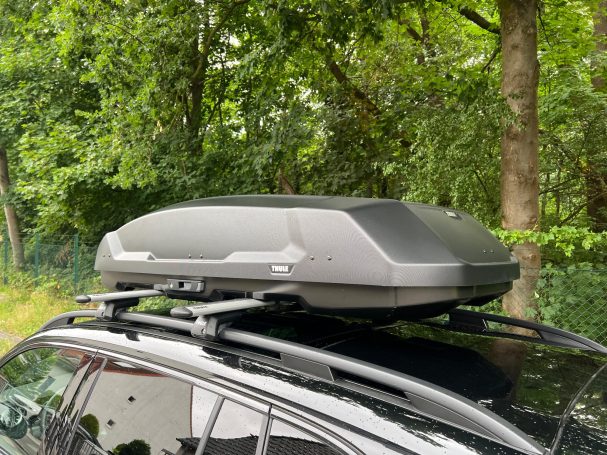 Dachbox auf einem Auto, platziert auf einem schwarzen Fahrzeugdach, umgeben von Bäumen.