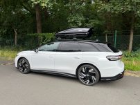 Weißer Kombi mit Dachbox steht an einem ruhigen Straßenrand, umgeben von Bäumen.