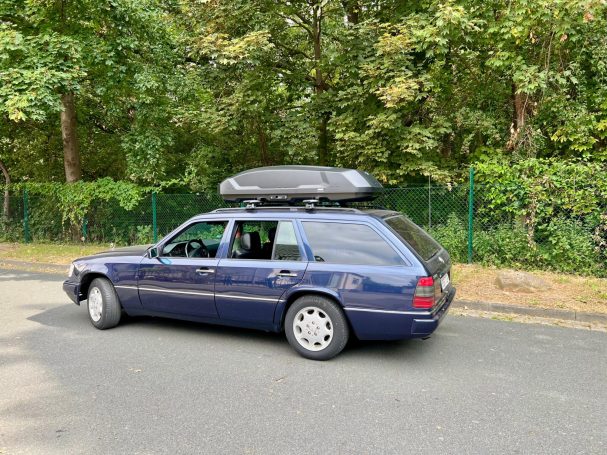 Blauer Kombi mit Dachbox parkt an einer Straße, umgeben von Bäumen.